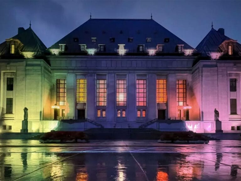 A large, illuminated building with colorful lights reflecting off wet pavement at dusk, featuring a symmetrical facade and multiple peaked roofs.