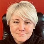 A person with short, light blonde hair sits in a black chair, wearing a dark turtleneck, against a red background.