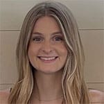 A person with long, light brown hair smiles at the camera in front of a light-colored wall.