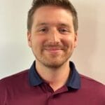 A man with short light brown hair and trimmed facial hair, wearing a maroon collared shirt, stands in front of a plain, light-colored wall, smiling slightly.