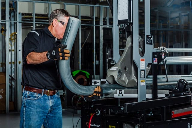 A man wearing safety glasses and gloves inspects a large metal pipe positioned on an industrial machine in a workshop setting.