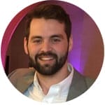 A man with short dark hair and a beard, wearing a light-colored jacket and white shirt, smiling at the camera against a blurred indoor background.