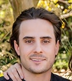 A young man with medium-length brown hair and light skin smiles at the camera outdoors, with greenery in the background.