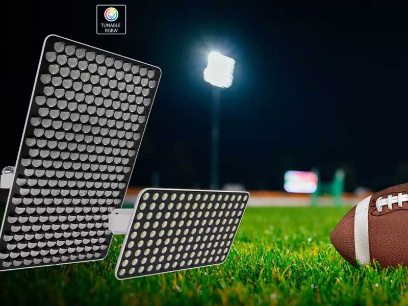 Two LED stadium lights are positioned on the grass near an American football, with an illuminated field in the background at night.